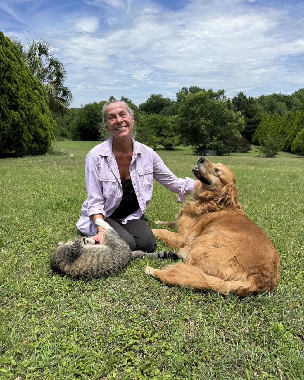 Vilena sitting in a sunny green field with her senior golden retriever and her tabby cat, all three relaxed and happy together